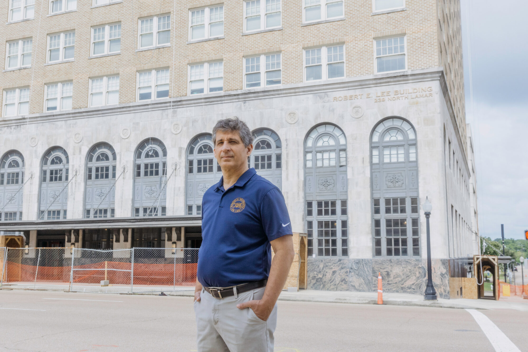 In a photograph, a White man with gray hair stands on the street in front of a concrete building with arched windows. The man is wearing a blue short-sleeved polo shirt with a logo.